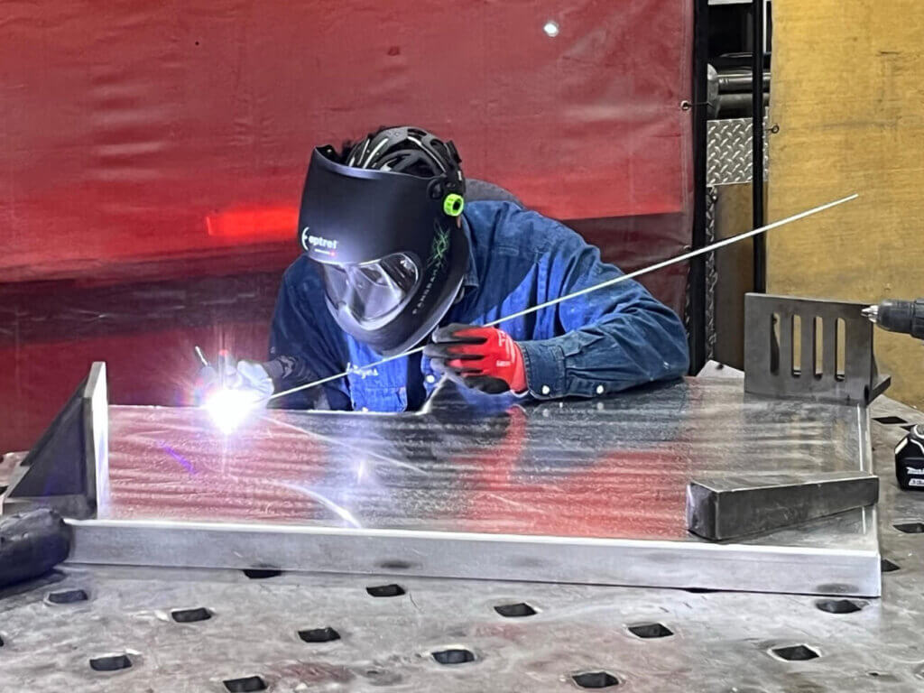 A welder wearing a protective helmet and gloves is welding a metal piece on a workbench